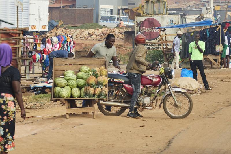 People on a motorcycle next to a wooden box full of watermelon and pineapple, Entebbe, Uganda