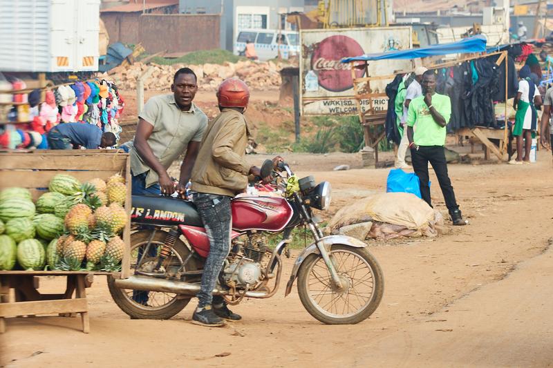 People on a motorcycle next to a wooden box full of watermelon and pineapple, Entebbe, Uganda