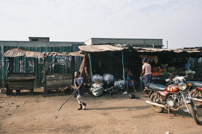 Child playing with a roller by a store front and fruit stand, Uganda